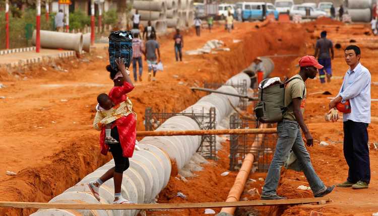A family in Angola balances over a narrow plank across a large Chinese construction site.